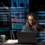 A woman data analyst is focused on her laptop in a dark office space, with the glow of SQL database code reflecting on the glass surface in front of her, symbolizing the process of transforming SQL query results into understandable visual formats such as tables and charts.